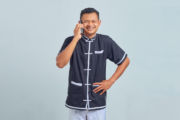 Retrato de jovem asiático feliz vestindo uniforme de caratê, falando no smartphone em fundo cinza