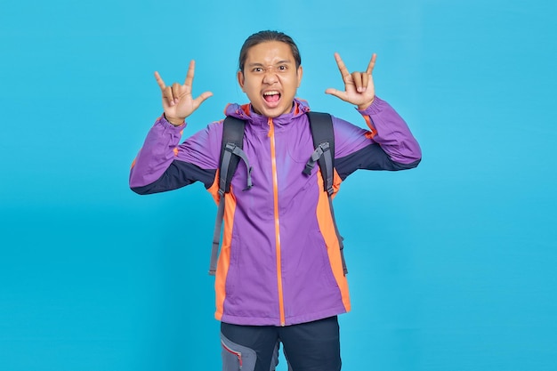 Retrato de jovem asiático gritando com uma expressão maluca fazendo o símbolo do rock com as mãos ao alto sobre fundo azul