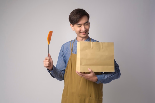 Retrato de jovem asiático usando avental segurando saco de papel e virador sobre estúdio de fundo branco