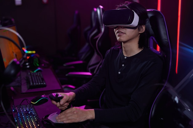 Retrato de jovem asiático usando fone de ouvido de RV enquanto joga videogame usando o racing shift no interior cibernético escuro, copie o espaço