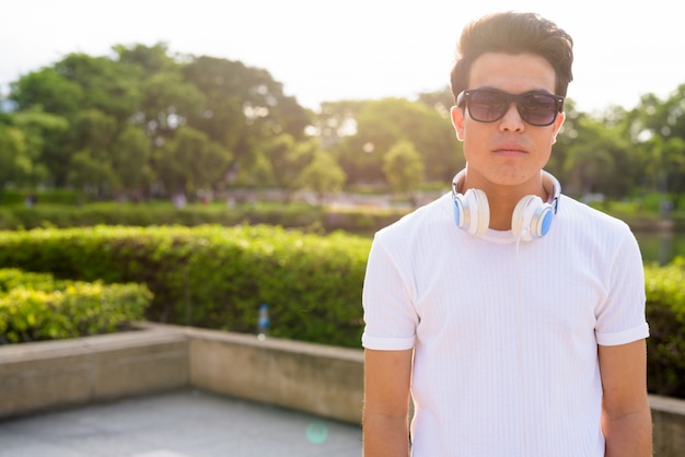 Retrato de jovem asiático usando fones de ouvido e óculos de sol enquanto relaxa no parque em Bangkok, Tailândia