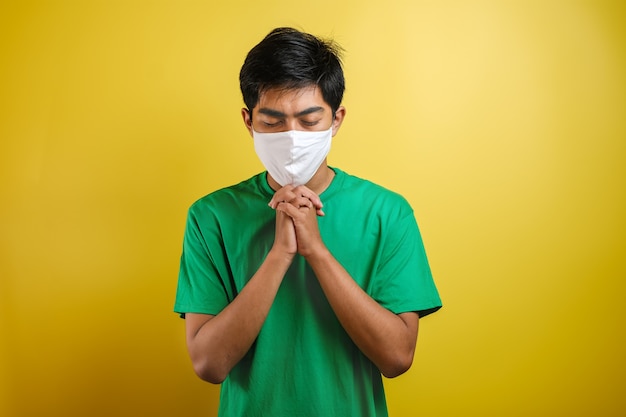 Retrato de jovem asiático usando máscara protetora contra o coronavírus, fazendo pray para o vírus corona é rapidamente superado isolado na cor amarela