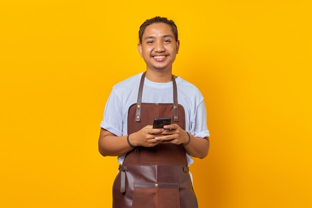 Retrato de jovem asiático vestindo avental e parecendo feliz usando o smartphone em fundo amarelo