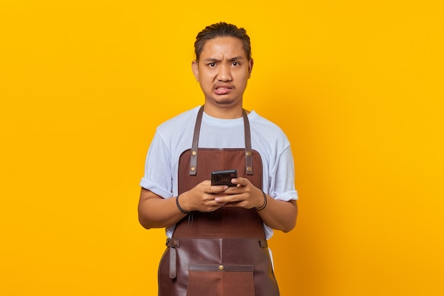 Retrato de jovem asiático vestindo avental, parecendo zangado e segurando o smartphone sobre fundo amarelo