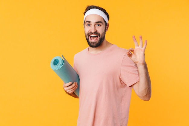Retrato de jovem atlético musculoso em camiseta sorrindo e carregando um tapete de fitness isolado na parede amarela