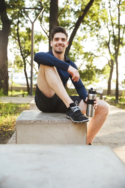Retrato de jovem Atlético vestido com roupas esportivas, sentado no banco e segurando uma garrafa de água enquanto faz exercícios no ensolarado parque verde