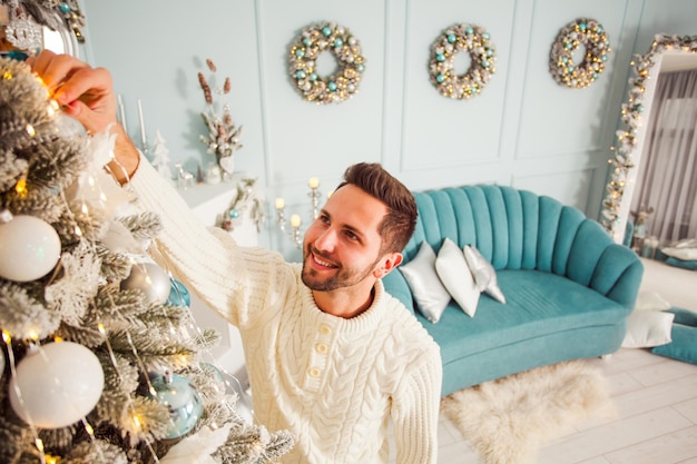 Retrato de jovem atraente e sorridente pendurado na decoração em uma árvore do abeto dentro de sua casa moderna e elegante Quarto ou estúdio com interior elegante na moda em cores azuis e brancas em um fundo