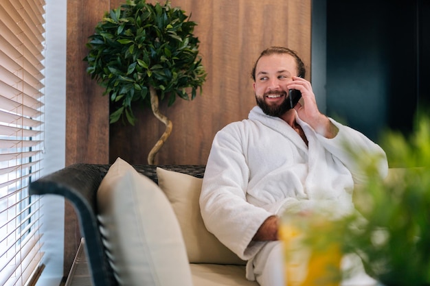 Retrato de jovem barbudo alegre vestindo roupão branco falando usando smartphone sorrindo olhando para longe sentado perto da janela no sofá confortável no salão spa após procedimentos de beleza