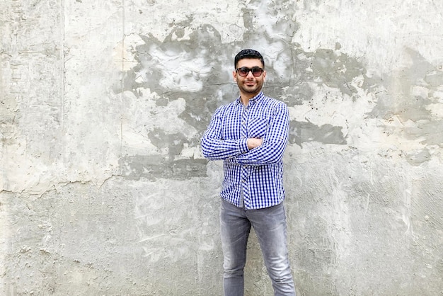 Retrato de jovem barbudo bem sucedido bonito com óculos de sol em pé de camisa azul quadriculada contra a parede de concreto cinza. braços cruzados e olhando para a câmera sorrindo.