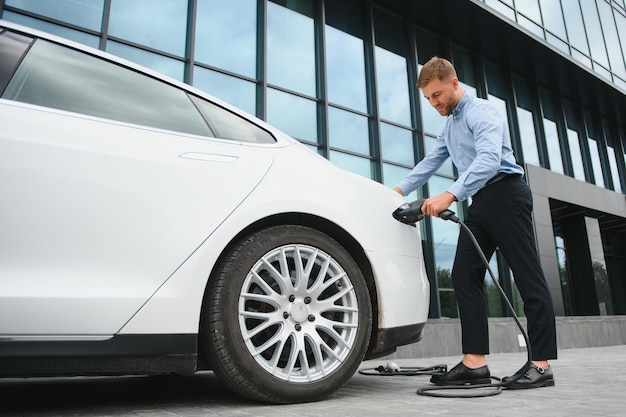 Retrato de jovem barbudo bonito em roupas casuais em pé na estação de carregamento e segurando um plugue do carregador para um carro elétrico Eco conceito de carro elétrico