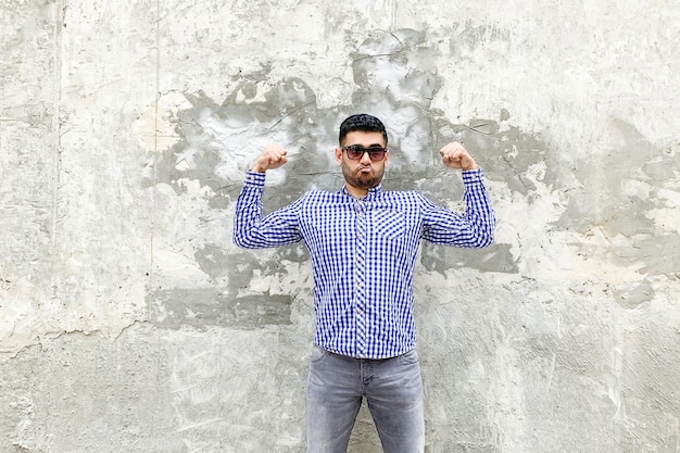 Retrato de jovem barbudo bonito orgulhoso na camisa azul quadriculada e óculos de sol em pé contra a parede cinza de concreto. olhando para a câmera em pose forte e mostrando que ele é forte e independente.