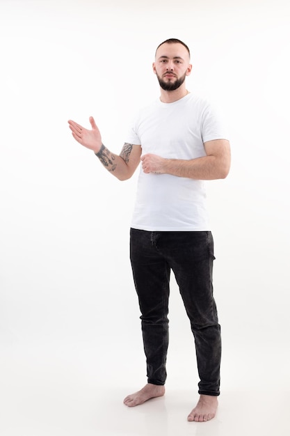 Retrato de jovem barbudo bonito sério, com cabelo escuro curto, vestindo jeans pretos, camiseta branca, levantando a mão com a palma da mão aberta apontando no fundo branco anúncio