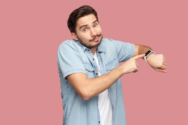 Retrato de jovem barbudo bonito triste em pé de camisa azul estilo casual apontando e mostrando seu relógio inteligente e olhando para a câmera. tiro de estúdio interno, isolado no fundo rosa.