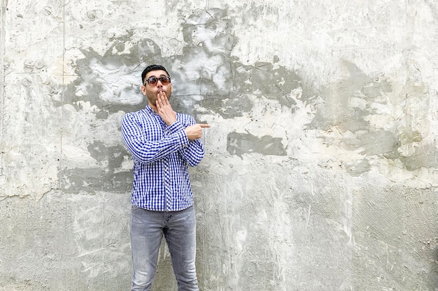 Retrato de jovem barbudo chocado em óculos de sol de camisa azul quadriculada em pé contra a parede cinza de concreto, apontando para o copyspace vazio de fundo e olhando para a câmera com cara de surpresa espantada