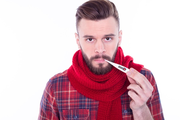 Retrato de jovem barbudo doente envolto em cachecol mede temperatura treme de frio fica em casa tem aparência doente tenta quebrar febre toma remédios Gripe sazonal Cuidados de saúde