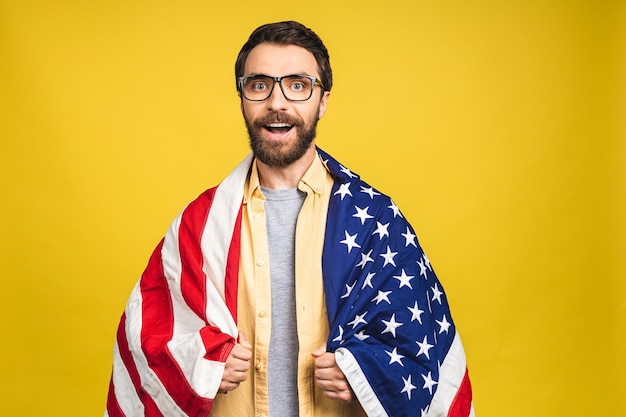 Retrato de jovem barbudo feliz segurando uma bandeira dos EUA isolada sobre fundo amarelo.