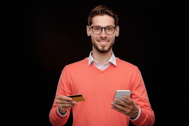 Retrato de jovem bonito alegre com a barba por fazer em pé com cartão de crédito e smartphone e usando o serviço de transação online