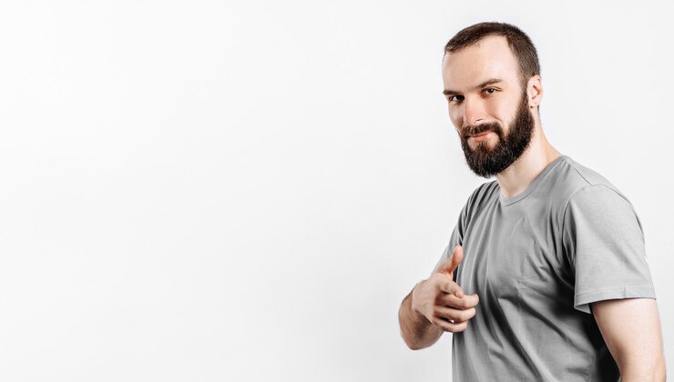 Foto retrato de jovem bonito alegre, sorrindo, olhando para a câmera, apontando o dedo para a frente sobre fundo branco, com espaço para simulação de publicidade