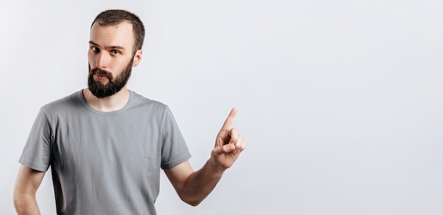 Retrato de jovem bonito alegre sorrindo olhando para a câmera apontando o dedo para cima em fundo branco com espaço para simulação de publicidade