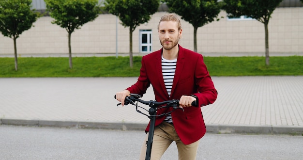 Retrato de jovem bonito caucasiano com casaco vermelho em pé ao ar livre com scooter elétrico e sorrindo para a câmera. Cara elegante e bonito no veículo na rua da cidade. Dolly shot. Ampliando.