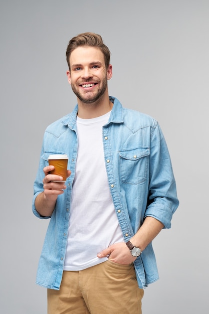 Retrato de jovem bonito com camisa jeans sobre a parede de luz segurando uma xícara de café para viagem