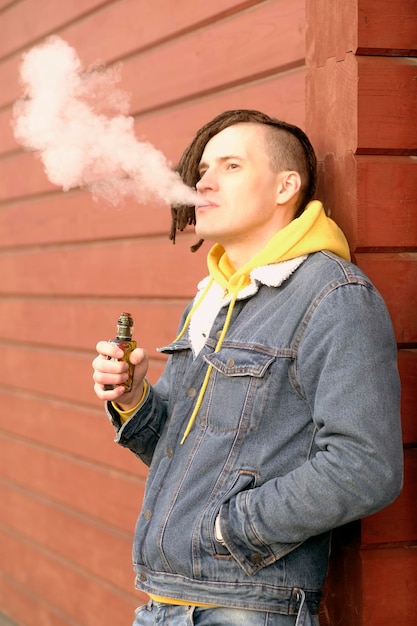 Retrato de jovem bonito com dreadlocks vaping em pé encostado na parede na rua da cidade Cara adulto fumando ecigarette soltando vapor