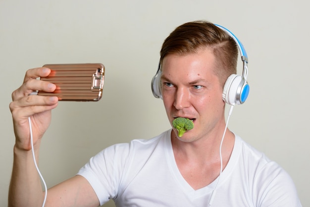 Retrato de jovem bonito com fones de ouvido, comendo brócolis e tomando selfie