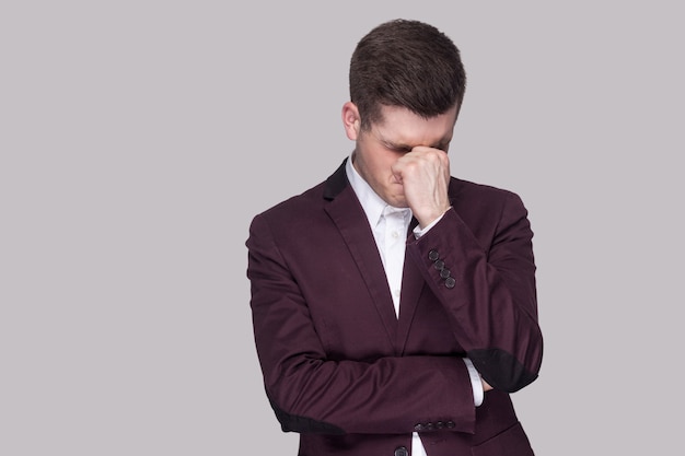 Retrato de jovem bonito deprimido triste em terno violeta e camisa branca, em pé, segurando a cabeça para baixo e as mãos no rosto e chorando. tiro de estúdio interno, isolado em fundo cinza.