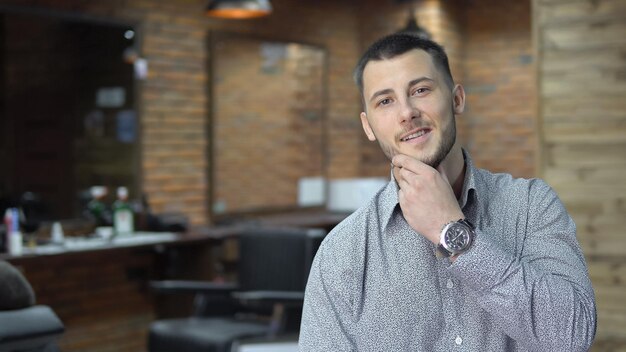 Retrato de jovem bonito e bem sucedido na barbearia após corte de cabelo no fundo do espelho desfocado e ferramentas de cabeleireiro