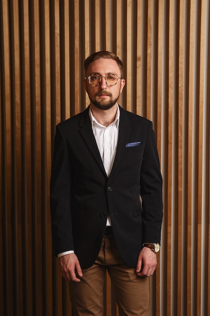 Retrato de jovem bonito e elegante de óculos, blazer e calças contra a parede de madeira