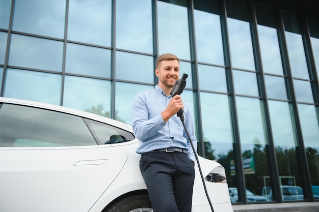 Retrato de jovem bonito em roupas casuais em pé no conceito de carro elétrico Eco da estação de carregamento