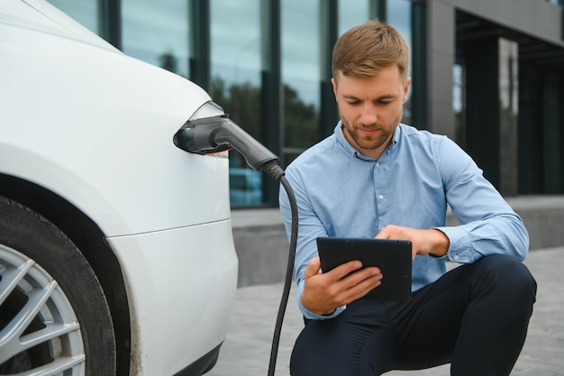 Retrato de jovem bonito em roupas casuais em pé no conceito de carro elétrico Eco da estação de carregamento