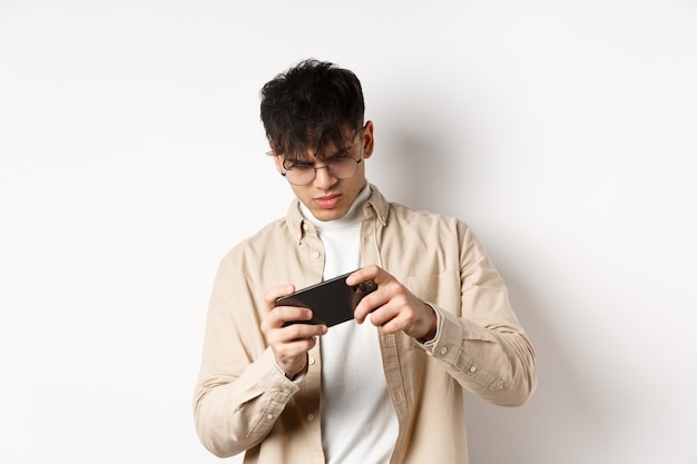 Foto retrato de jovem bonito jogando videogame no celular, incline o smartphone para jogar corridas, de pé na parede branca.