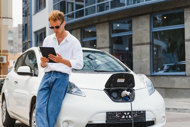 Retrato de jovem bonito no vestuário desportivo, de pé na estação de carregamento. Conceito de carro elétrico ecológico