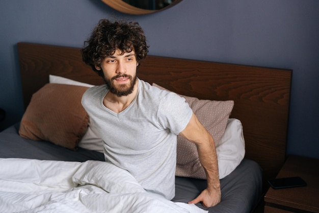 Retrato de jovem bonito sentado na cama depois de acordar e dar as boas-vindas à manhã olhando pela janela