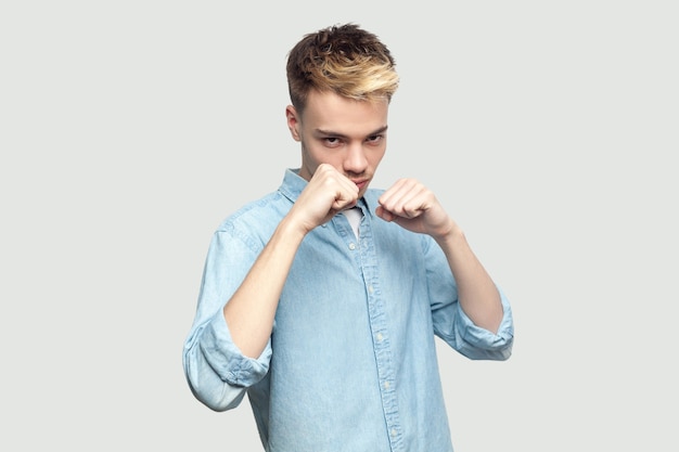 Retrato de jovem bonito sério na camisa azul clara em pé em punhos de boxe, olhando para a câmera e pronto para atacar ou defesa. estúdio interno atirou no espaço da cópia de fundo cinza.