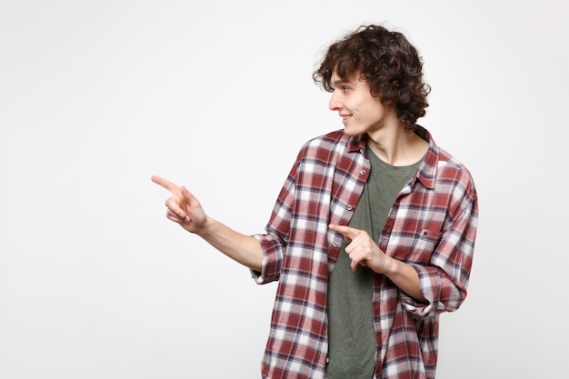 Retrato de jovem bonito sorridente em roupas casuais, olhando, apontando o dedo indicador de lado, isolado no fundo branco no estúdio. Emoções sinceras de pessoas, conceito de estilo de vida. Simule o espaço da cópia.