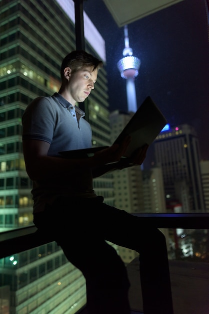 Retrato de jovem bonito usando laptop contra uma janela de vidro com vista da cidade à noite