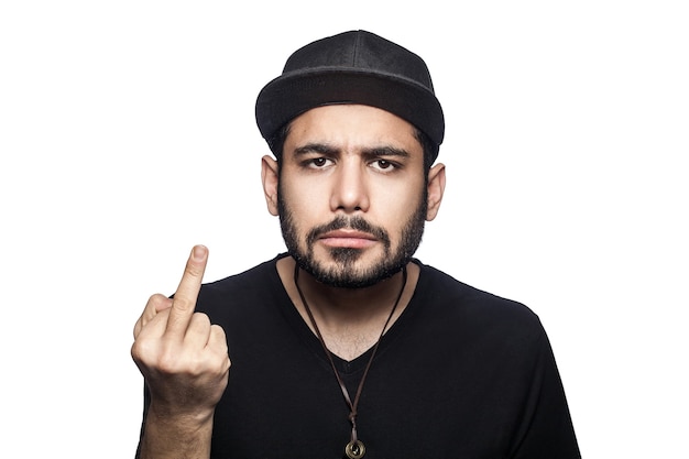 Retrato de jovem bravo com camiseta preta e boné, olhando para a câmera e mostrando o dedo médio. tiro do estúdio, isolado no fundo branco.