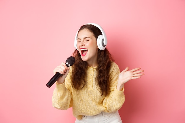 Retrato de jovem cantando karaokê, usando fones de ouvido e cantando, segurando o microfone, em pé contra a parede rosa.