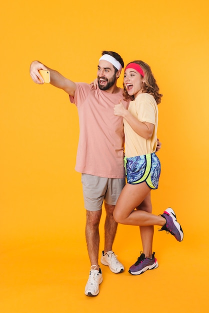 Retrato de jovem casal atlético usando bandanas, sorrindo e tirando uma foto de selfie no celular isolada sobre a parede amarela