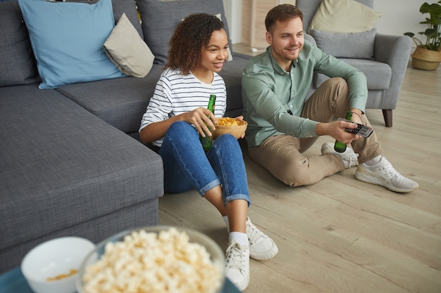 Retrato de jovem casal de raça mista assistindo TV em casa e bebendo cerveja enquanto está sentado no chão de um apartamento aconchegante