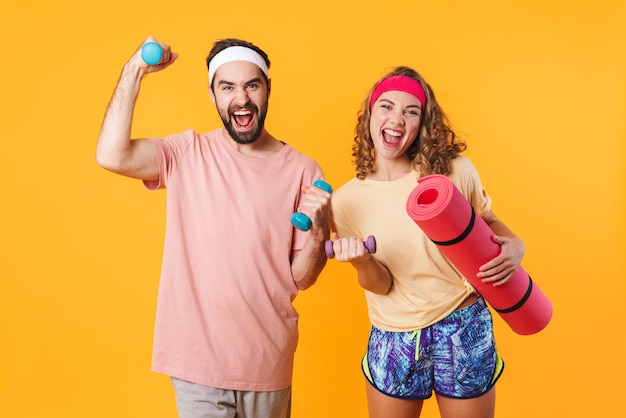 Retrato de jovem casal feliz atlético usando bandanas segurando halteres e um tapete de fitness isolado sobre a parede amarela