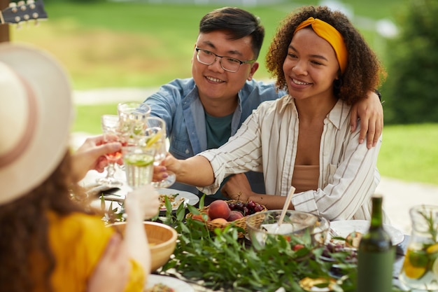 Retrato de jovem casal mestiço brindando enquanto janta com amigos ao ar livre na festa de verão