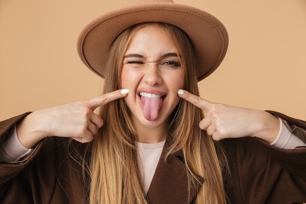 Retrato de jovem caucasiana com chapéu e casaco se divertindo e mostrando a língua isolada em bege