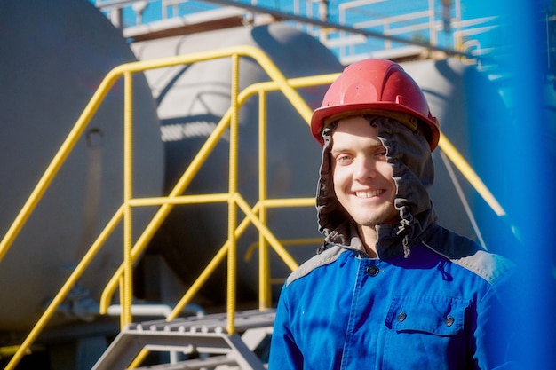 Retrato de jovem caucasiano em capacete de construção e roupas de trabalho contra o fundo de grandes cisternas no dia de verão Trabalhador olha para a câmera e sorri Pessoa positiva