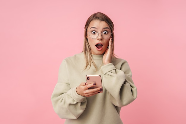 Retrato de jovem chocado com óculos usando o celular e olhando para a câmera, isolado sobre a parede rosa