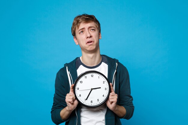 Retrato de jovem chorando cansado em roupas casuais, segurando o relógio redondo isolado na parede azul. o tempo está se esgotando. conceito de estilo de vida de emoções sinceras de pessoas.