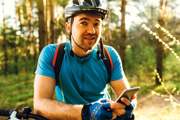 Retrato de jovem ciclista caucasiano em pé na floresta segurando o telefone celular usando o aplicativo on-line para pesquisar coordenadas GPS enquanto andava de bicicleta Esporte de viagem e conceito de tecnologia moderna