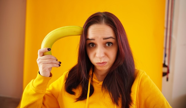 Foto retrato de jovem com banana em fundo amarelo close-up de fêmea com capuz amarelo brinca com frutas imaginando-o como arma